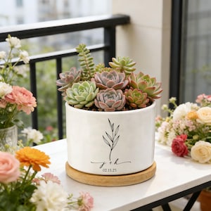 May include: A white ceramic planter filled with assorted succulents, resting on a round wooden base. The planter features a black botanical design and the initials "j + h" with the date "02.15.25" in a delicate script. The arrangement is set on a white surface.