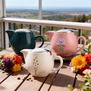 Op de afbeelding: Drie keramische theepotten in teal, roze en crème, elk met een roestvrijstalen deksel en een handvat. De theepotten zijn versierd met bloemmotieven en de naam "Rachel". De theepotten staan op een houten tafel met bloemen.