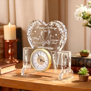 May include: A clear crystal clock with a heart-shaped top, featuring the words "Forever & Always" and "Love." The clock face is gold-toned with black Roman numerals. The base spells out "LOVE."