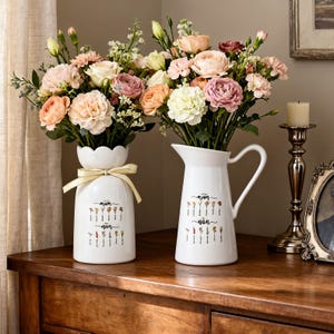 May include: Two white ceramic vases, each holding a bouquet of flowers, are displayed on a wooden surface. The vase on the left has a scalloped rim and a yellow ribbon. Both vases feature floral illustrations and the words "Mum" and "Nan".