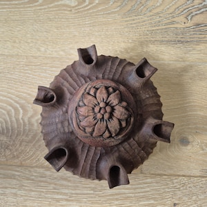 May include: A dark brown, hand-carved wooden flower pot featuring a central floral design. The circular pot has multiple openings around the edge. The wood has a textured finish, and is a rich, dark brown colour.