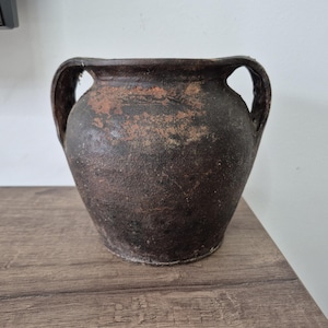 May include: A dark brown, aged ceramic vase with two handles. The vase has a rounded body and a wide opening. The surface has a textured, weathered appearance, with variations in color. The vase is sitting on a wooden surface.