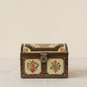 May include: A rectangular metal chest with a hinged lid and a central metal latch. The exterior features a wood-grain pattern with cream-coloured panels decorated with colourful floral arrangements in vases and red heart motifs on the front and lid.