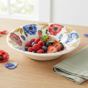 May include: A white ceramic bowl featuring a painted floral pattern of red and blue poppies. The bowl is filled with a mixture of fresh raspberries, blueberries, and sliced strawberries, garnished with a small sprig of green mint leaves.
