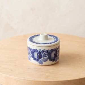 Puede incluir: Un pequeño tarro cilíndrico de cerámica con tapa a juego, que presenta un patrón floral azul pintado a mano sobre un fondo blanco. La tapa tiene un pomo central y un borde festoneado azul, mientras que el cuerpo muestra flores azules.