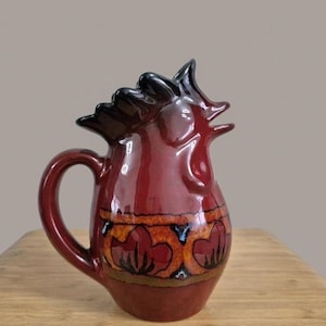 Può includere: Una brocca in ceramica color bordeaux con una parte superiore e un manico a forma di gallo. La brocca presenta una fascia decorativa di cuori e motivi floreali in arancione, marrone e nero. La brocca è su una superficie di legno.