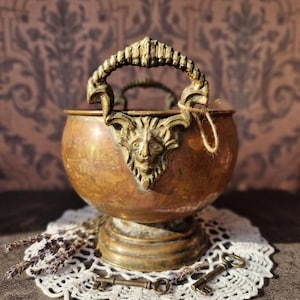 Può includere: Una ciotola rotonda in rame con base a piedistallo e due manici in ottone decorati con un motivo a testa di leone. La ciotola presenta una patina invecchiata ed è accompagnata da due piccole chiavi in metallo e un rametto di lavanda essiccata su un centrino di pizzo.