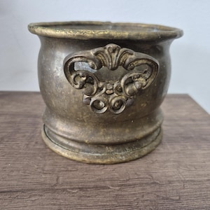 Op de afbeelding: Een vintage messing container met een sierlijke handgreep. De container heeft een afgeronde vorm met een brede opening en een decoratieve handgreep. Het messing heeft een aangetaste, verouderde uitstraling, wat suggereert dat het een antiek is. De container staat op een houten oppervlak.