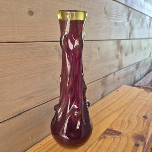 Puede incluir: Un jarrón de vidrio alto y esbelto con un cuerpo de color rojo burdeos profundo y una superficie orgánica texturizada. El jarrón tiene un cuello estrecho que se ensancha ligeramente en la parte superior, rematado con una banda de vidrio verde lima translúcido.