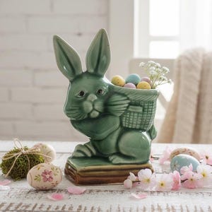 Könnte beinhalten: Ein grüner Keramik-Hasen-Pflanztopf mit einem Korb auf dem Rücken, gefüllt mit bunten Ostereiern und weißen Blumen. Der Hase sitzt auf einem braunen Sockel. Dekorative Ostereier und rosa Blütenblätter sind um den Pflanztopf verstreut.