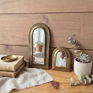 Pode incluir: Dois espelhos arqueados com molduras de latão envelhecido estão posicionados na vertical. O espelho mais alto está à esquerda do mais curto. Ambos os espelhos possuem uma superfície de vidro refletora numa moldura metálica dourada.