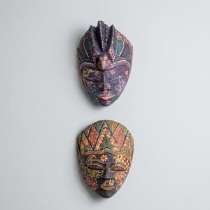 Può includere: Due maschere in legno intagliate a mano con dettagli dipinti complessi. La maschera superiore è prevalentemente viola con motivi floreali e una cresta centrale. Quella inferiore presenta un design geometrico e floreale multicolore in verde, arancione e marrone.