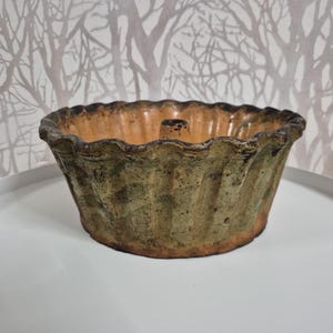 Puede incluir: Un molde para bizcocho de cerámica acanalado y rústico con un borde festoneado. El molde presenta un esmalte moteado verde y marrón con un interior naranja. El molde está sobre una superficie blanca, con un fondo estampado.