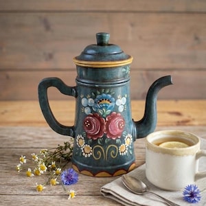 Op de afbeelding: Een handbeschilderde, blauwgroene koffiepot met een bloemenontwerp. Een witte mok met een schijfje citroen en een zilveren lepel liggen op een houten oppervlak, samen met wilde bloemen. De koffiepot is ongeveer 20 cm hoog.