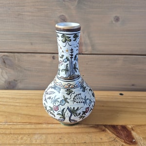 Könnte beinhalten: Eine weiße Keramikvase mit bauchigem Körper und schlankem Hals. Die Vase ist mit einem Blumenmuster in Blau, Grün und Rot verziert. Der Rand ist braun und der Boden grün.
