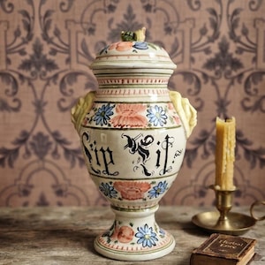 Op de afbeelding: Een crèmekleurige keramische apothekerspot met deksel, voorzien van handgeschilderde blauwe en oranje bloemmotieven, gele handvatten aan de zijkanten en de zwarte tekst "Sip It" in een decoratief lettertype op het midden van de pot.