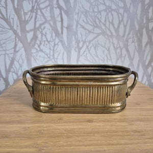 Op de afbeelding: Een ovale koperen plantenbak met een geribbelde buitenkant en twee kleine handvatten aan de zijkanten. Het metaal heeft een warme, verouderde patina met zichtbare horizontale banden langs de boven- en onderranden van de bak.