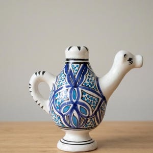 Può includere: Brocca in ceramica bianca con beccuccio e manico, ornata da motivi floreali blu e turchese. La brocca ha una base a piedistallo e una parte superiore decorativa con dettagli neri. Il design ricorda la ceramica tradizionale.