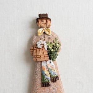 Pode incluir: Estatueta de cerâmica de uma pessoa usando chapéu, casaco e cachecol marrons. A estatueta está segurando um balde de madeira e um maço de folhas verdes. O cachecol tem um padrão floral colorido. A estatueta tem um rosto sorridente.