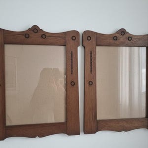 Op de afbeelding: Twee bijpassende houten fotolijsten met een donkerbruine afwerking. Elke lijst heeft een rechthoekige opening, decoratieve details en een gebogen bovenkant. De lijsten worden tegen een witte muur getoond.