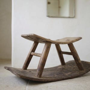 Puede incluir: Un taburete mecedora de madera pequeño y bajo con un asiento curvo en forma de silla de montar, sostenido por cuatro patas anguladas conectadas con travesaños horizontales. Toda la pieza está hecha de madera marrón oscura desgastada.