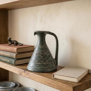 Pode incluir: Uma prateleira de madeira com um jarro verde escuro padronizado com uma alça, uma pequena figura de raposa de bronze e uma pilha de livros. Os livros têm títulos como "The Silent Forest" e "Ancient Weaves". Um pequeno livro de cor clara também está presente.
