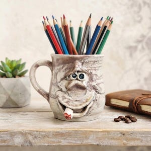 Puede incluir: Una taza de cerámica con un esmalte texturizado y agrietado de color blanco roto, que presenta un rostro esculpido con ojos grandes y una boca ancha que sostiene un pequeño objeto cilíndrico. La taza está llena de lápices de colores rojos, azules, verdes y marrones.