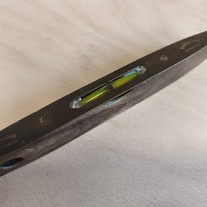 May include: A dark metal spirit level featuring a central glass vial filled with yellow-green liquid. The tool is engraved with the text C.F. JOHNSON SHEFFIELD and WARRANTED CORRECT. The body is elongated with a tapered, pointed end design.