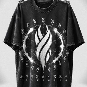 May include: A black short-sleeve t-shirt with a distressed wash finish. The front features a large white graphic of a central spiral marker symbol surrounded by a circular ring of letters A through Z and a bottom row of numbers 1 through 0.