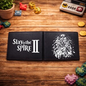 Op de afbeelding: Zwart dubbelgevouwen portemonnee met de tekst "Slay the Spire II" in het wit. De rechterkant van de portemonnee toont een gedetailleerde witte afbeelding van personages. De portemonnee is open en toont een bruin interieur en stiksels.