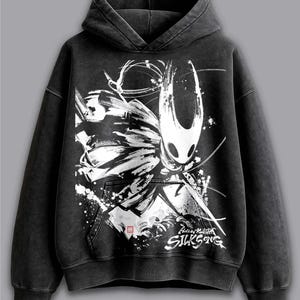 May include: Black hooded sweatshirt with a white and black graphic of a Hollow Knight character. The design includes the text "Hollow Knight Silksong". The sweatshirt has a kangaroo pocket and a drawstring hood.