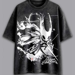 May include: Dark grey t-shirt with a white and black graphic of a character from the video game Hollow Knight. The graphic includes the text "Hollow Knight SILKSONG". The shirt has a vintage, washed appearance.