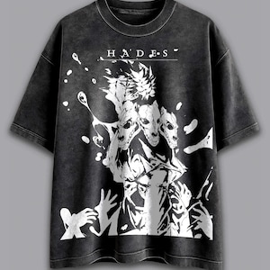Può includere: T-shirt grigio scuro con finitura invecchiata. Il davanti presenta una grafica bianca di una creatura a tre teste e la scritta "HADES". Il design include forme astratte e figure simili a mani.