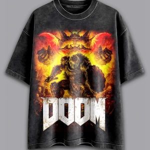 May include: Dark gray t-shirt featuring a detailed graphic from the video game "Doom." The design includes a warrior figure with a shield, set against a fiery background, with the word "DOOM" in bold, white letters at the bottom.