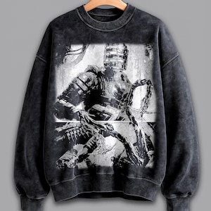 May include: A dark gray sweatshirt featuring a black and white graphic of a futuristic armored figure holding a chain. The image has a distressed, vintage look. The sweatshirt has a crew neck and long sleeves.