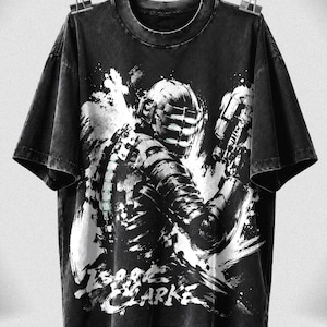 May include: A black short-sleeve t-shirt featuring a distressed graphic print of a character in a futuristic helmet and suit holding a weapon. The design includes white paint-splatter effects and the text Isaac Clarke printed in a bold, stylized font.