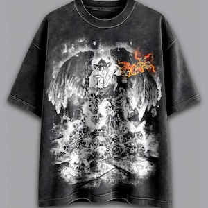 May include: Dark grey t-shirt with a graphic of a winged figure, skulls, and flames in white, black, and orange. The tee has a distressed, vintage appearance. The design is centered on the front of the shirt.