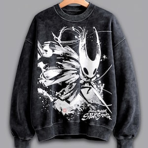 May include: Black sweatshirt with a white and black graphic of a character from the video game Hollow Knight. The graphic includes the text "Hollow Knight Silksong". The sweatshirt has a crew neck and long sleeves.