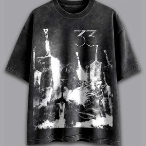 以下が含まれることがあります： 色あせたグラフィックプリントのダークグレーTシャツ。デザインは、様式化された剣と数字の「33」を白で表現しています。Tシャツはクルーネックと半袖です。