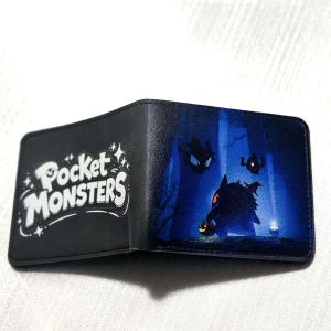 May include: Black bi-fold wallet featuring the text "Pocket Monsters" in white. The interior design showcases a spooky forest scene with cartoon characters and a jack-o'-lantern. The wallet is made of a smooth, dark material.