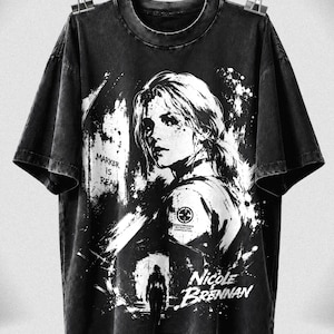 May include: A black acid-wash t-shirt featuring a white graphic print of a person with the text "MARKER IS REAL" and "NICOLE BRENNAN". The design includes a small circular logo on the shoulder and a silhouette figure in the lower section.