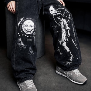 Può includere: Pantaloni in denim nero lavaggio acido con stampe grafiche bianche. La gamba destra mostra un design a faccia circolare con il testo "TERMINA" sotto. La gamba sinistra presenta una figura in piedi all'interno di una cornice circolare e coulisse.