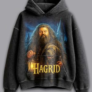 May include: Black hooded sweatshirt featuring a portrait of Hagrid from the Harry Potter series, holding a lantern and accompanied by a dog. The word "HAGRID" is printed in gold on the front pocket. The background shows a castle under a night sky.