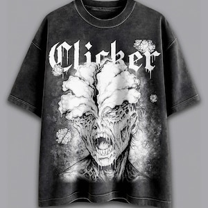 Puede incluir: Camiseta gris oscuro con una ilustración en blanco y negro de una cara de zombi y la palabra "Clicker" en fuente gótica. El diseño incluye crecimientos fúngicos y una textura desgastada.