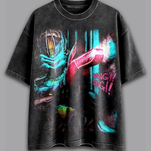 May include: Dark gray t-shirt featuring a neon illustration of a futuristic figure holding a weapon. The design includes blue, red, and yellow accents, with the text "BANG!!" in a comic book style. The shirt has a vintage, washed look.