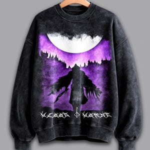 May include: Black and grey sweatshirt with a purple and white graphic. The graphic features a silhouetted figure with outstretched arms, a crescent moon, and text at the bottom. The sweatshirt is hanging on a wooden hanger.