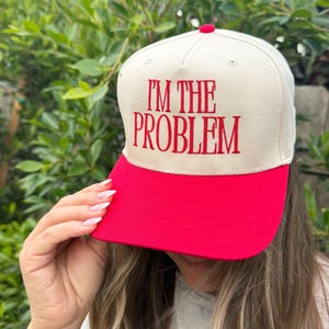 Peut inclure: Casquette de baseball beige et rouge avec le texte brodé "I'M THE PROBLEM". La casquette a une visière rouge et un bouton rouge sur le dessus. Le chapeau est de style camionneur classique.