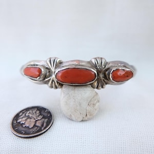 Puede incluir: Un brazalete plateado con tres piedras ovaladas de coral rojo. El brazalete tiene detalles decorativos con forma de flor. Una moneda de plata está al lado del brazalete. El brazalete se exhibe sobre una pequeña piedra blanca.