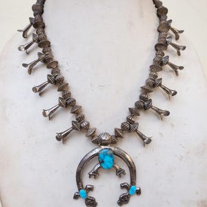 Puede incluir: Un collar de plata con diseño de flor de calabaza y detalles turquesa. El collar presenta una serie de flores de plata y un colgante central con una piedra turquesa. El colgante tiene forma de herradura.