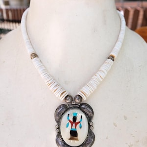 Pode incluir: Um colar com um fio de contas brancas em forma de disco e um pendente central de prata. O pendente tem uma incrustação oval que representa uma figura estilizada em vermelho, preto e turquesa, emoldurada por metal prateado decorativo.